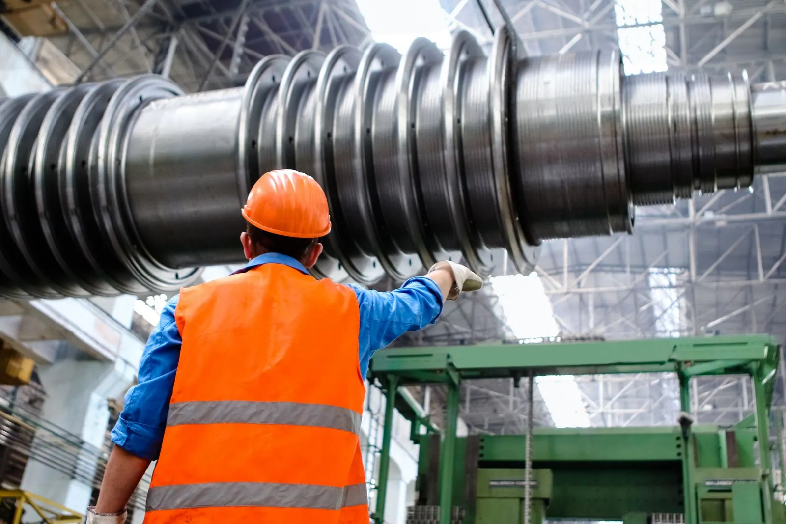 Industriearbeiter mit Schutzhelm und Warnweste kontrolliert eine große Anlagenwelle in einer Werkhalle